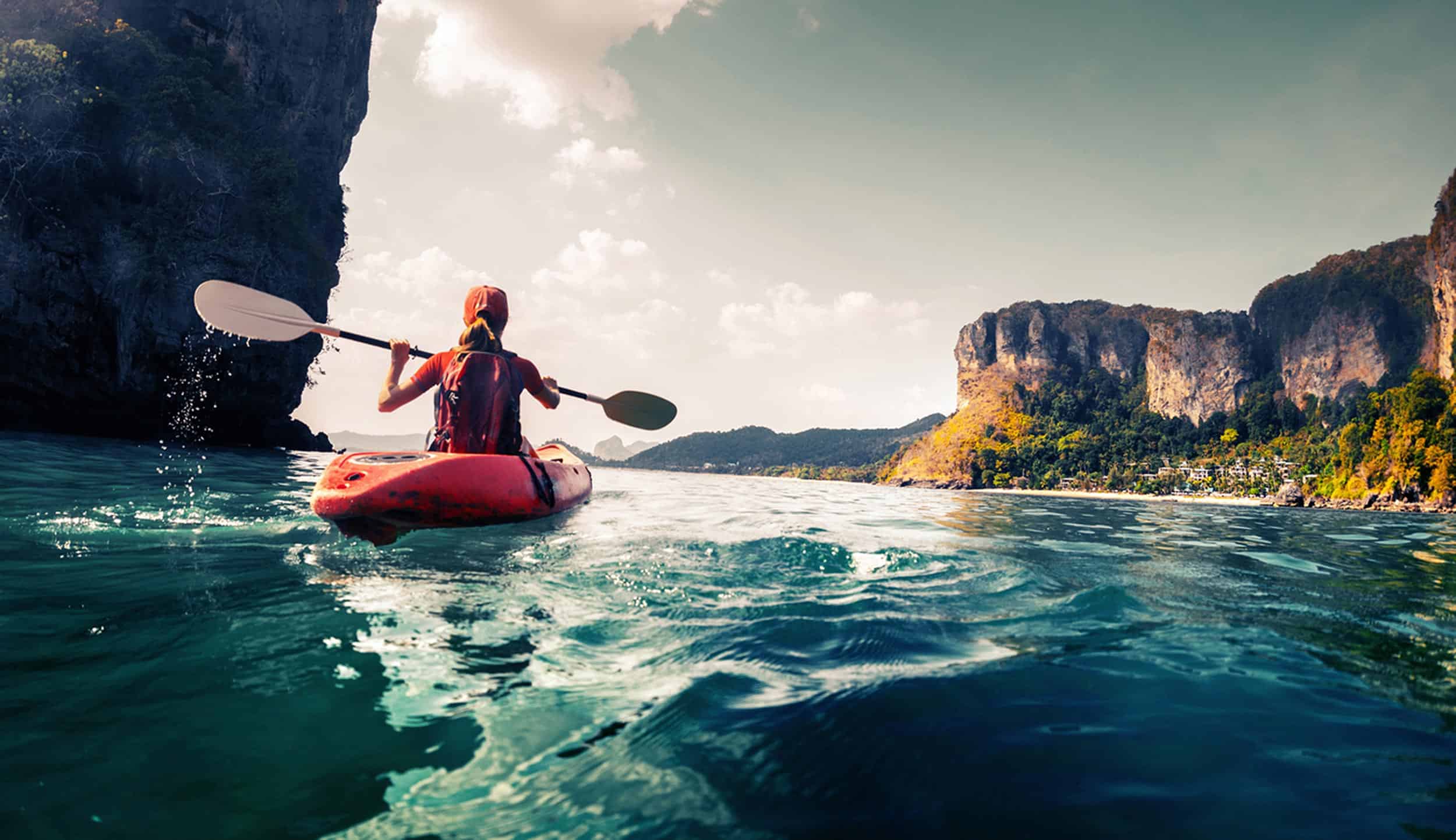 Woman kayaking on blue body of water between cliffs toward the setting sun